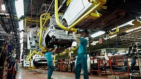 The image depicts human workers assembling a vehicle on an automotive production line