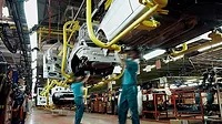 The image depicts human workers assembling a vehicle on an automotive production line