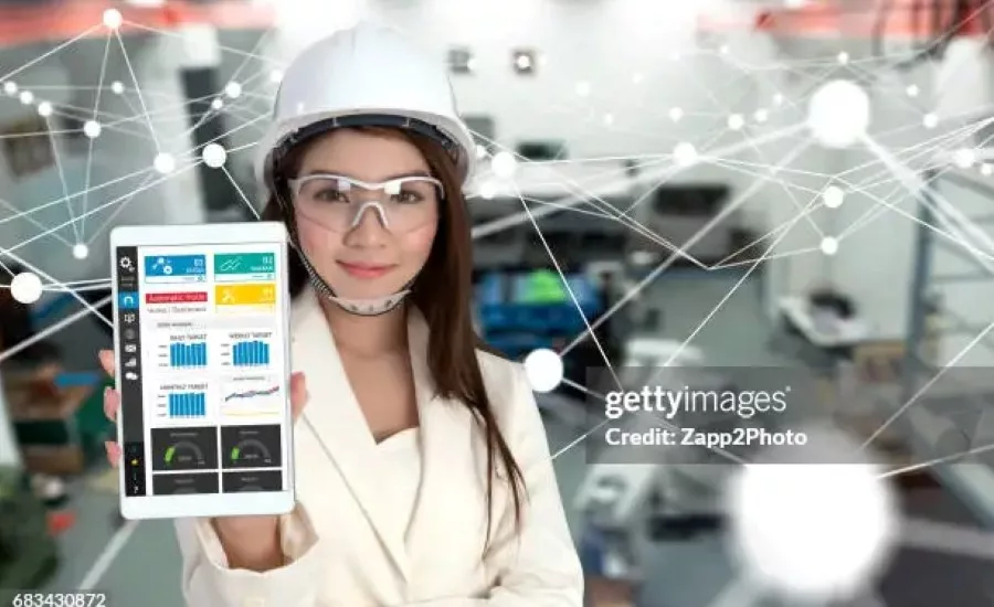 Engineer wearing safety glasses and a hard hat displaying a factory analytics dashboard on a tablet, illustrating connected manufacturing and industrial IoT technologies.