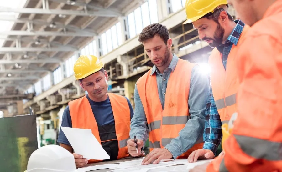 Manufacturing engineers and plant workers wearing hard hats and safety vests reviewing production plans and project documents on a factory floor.