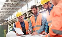 Manufacturing engineers and plant workers wearing hard hats and safety vests reviewing production plans and project documents on a factory floor.