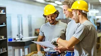 Manufacturing Team Reviewing Production Plans Manufacturing engineers and plant workers wearing hard hats and safety vests reviewing production plans and project documents on a factory floor.