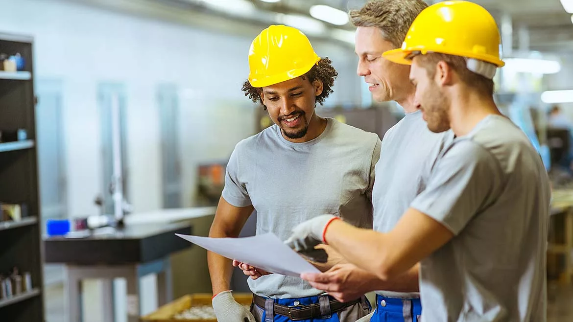 Manufacturing engineers and plant workers wearing hard hats and safety vests reviewing production plans and project documents on a factory floor.