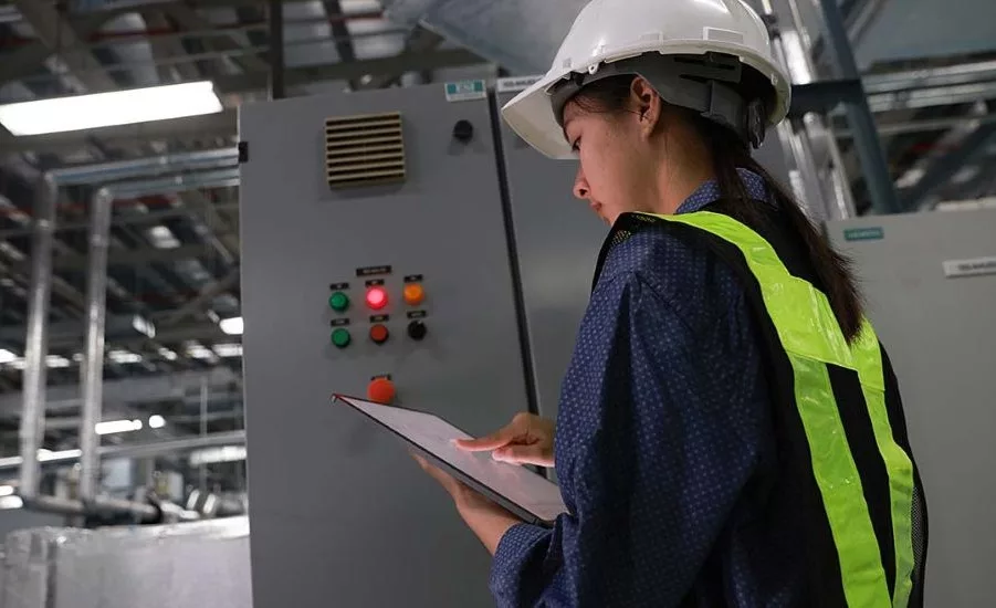 Industrial worker wearing a hard hat and high-visibility vest inspecting equipment and reviewing maintenance data on a tablet in a manufacturing facility.