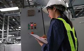 Industrial worker wearing a hard hat and high-visibility vest inspecting equipment and reviewing maintenance data on a tablet in a manufacturing facility.