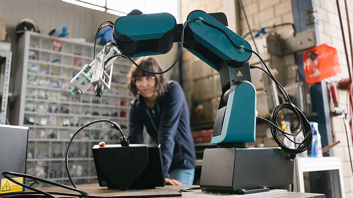 Collaborative robot equipped with a precision tool operating in a manufacturing workspace while an engineer monitors the robotic system in the background.