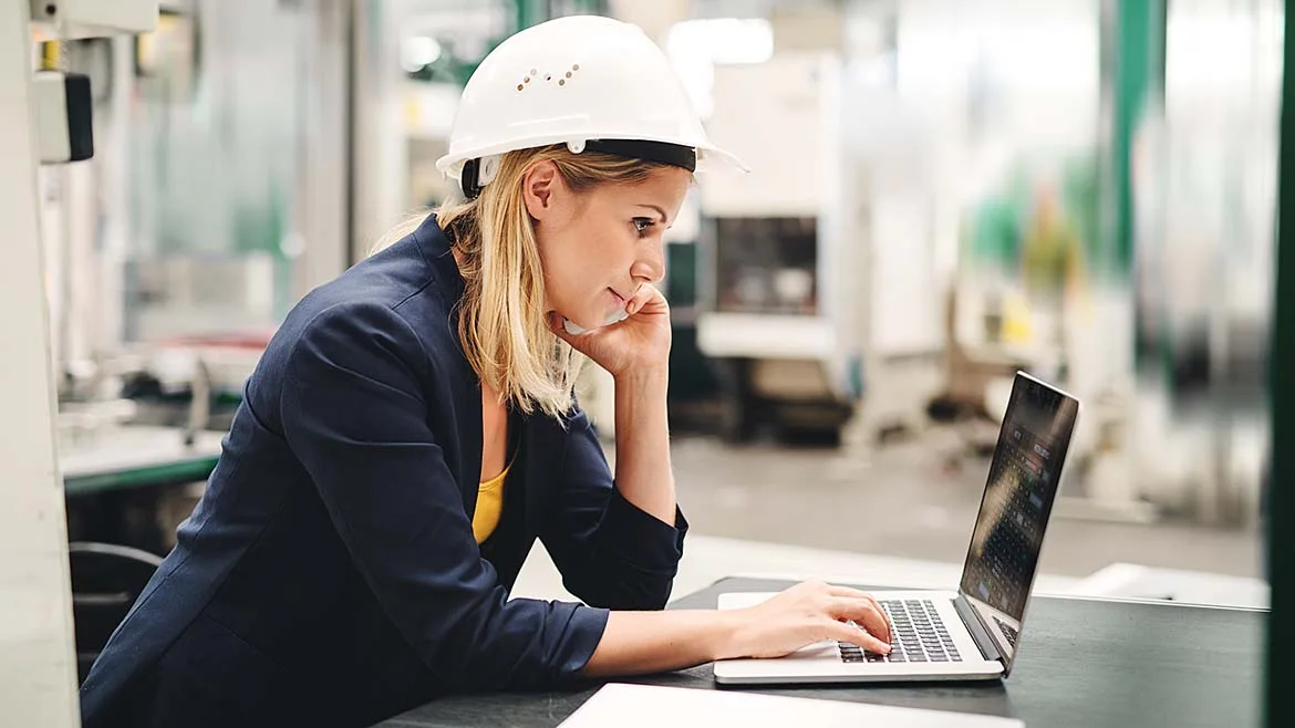 Engineer Working on Laptop in Manufacturing Facility Engineer wearing a hard hat using a laptop in a manufacturing facility.