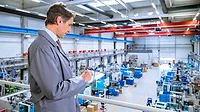Manufacturing Manager Monitoring Factory Operations Manufacturing manager reviewing production data on a tablet overlooking a factory floor.