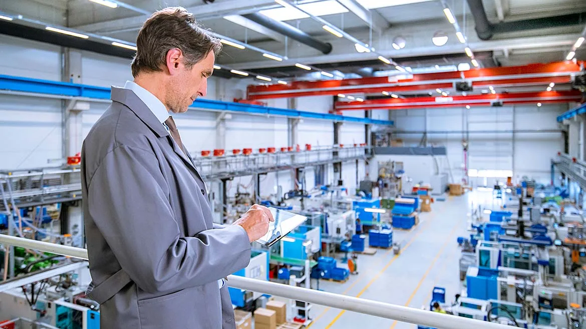 Manufacturing manager reviewing production data on a tablet overlooking a factory floor.