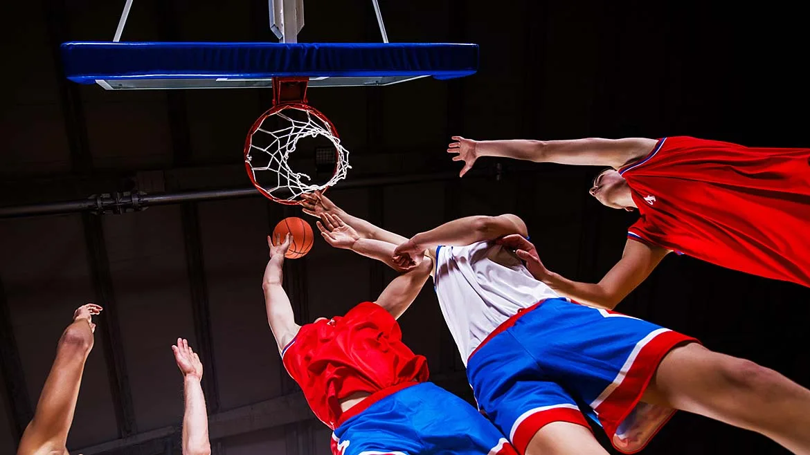 Basketball players reaching for the ball beneath the hoop during a game.