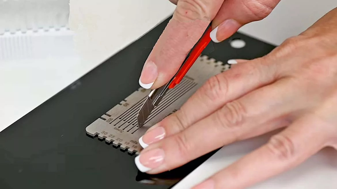 Close-up of a person using a utility knife and a Temper II film applicator gauge on a coated metal panel to measure coating thickness and evaluate film application quality.