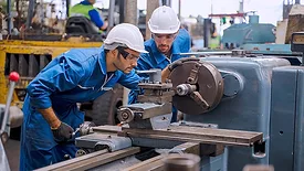 Two factory workers in blue overalls and white safety hats and safety eyewear, working in at factory.