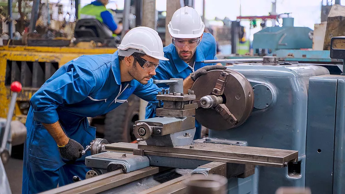 Two factory workers in blue overalls and white safety hats and safety eyewear, working in at factory.