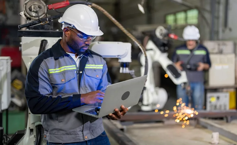 A worker wearing a work jacket, safety glasses and white hard hat working on a laptop in a factory. A robotic arm and worker is blurred in the background.