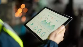 Cropped image of person in a safety vest, holding a laptop with analytics on the screen and a blurred background.