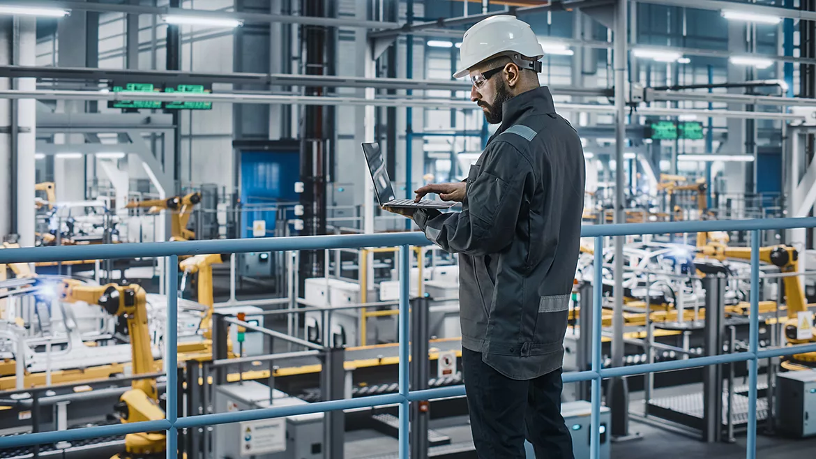 Car factory engineer in work uniform and white safety hat using a laptop.