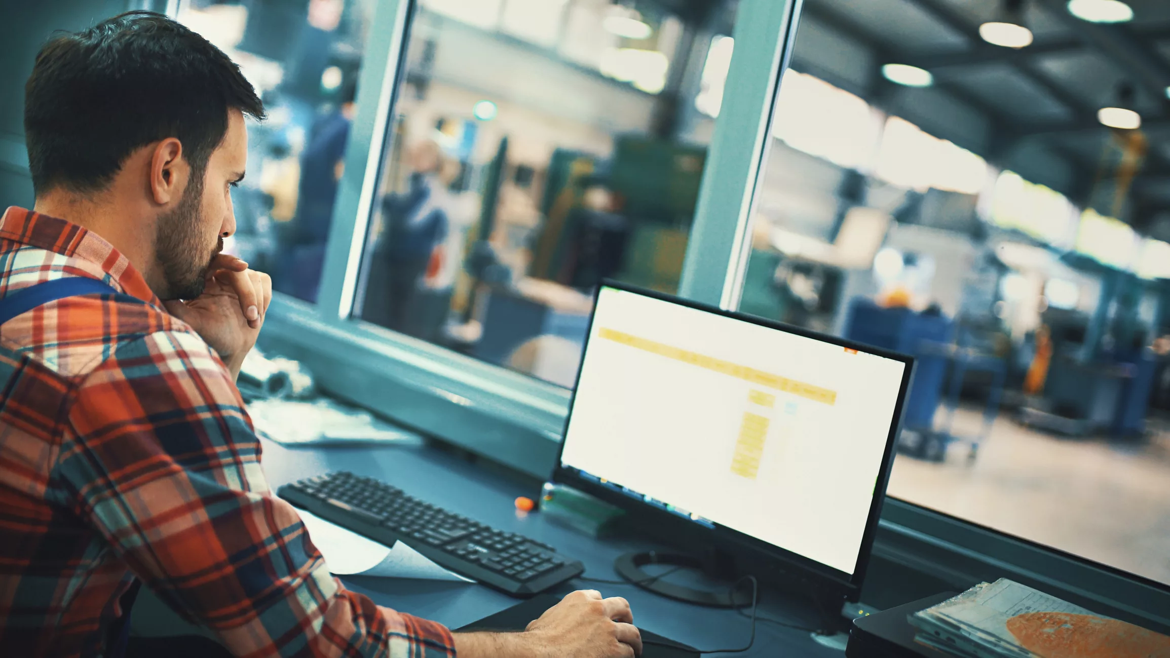 Man in plaid long-sleeve shirt, working on the computer, a blurred factory floor in the background.