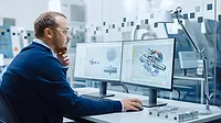 Male industrial engineer working on a personal computer with two monitor screens. The monitors show CAD software with 3D prototype of hybrid electric engine being tested.