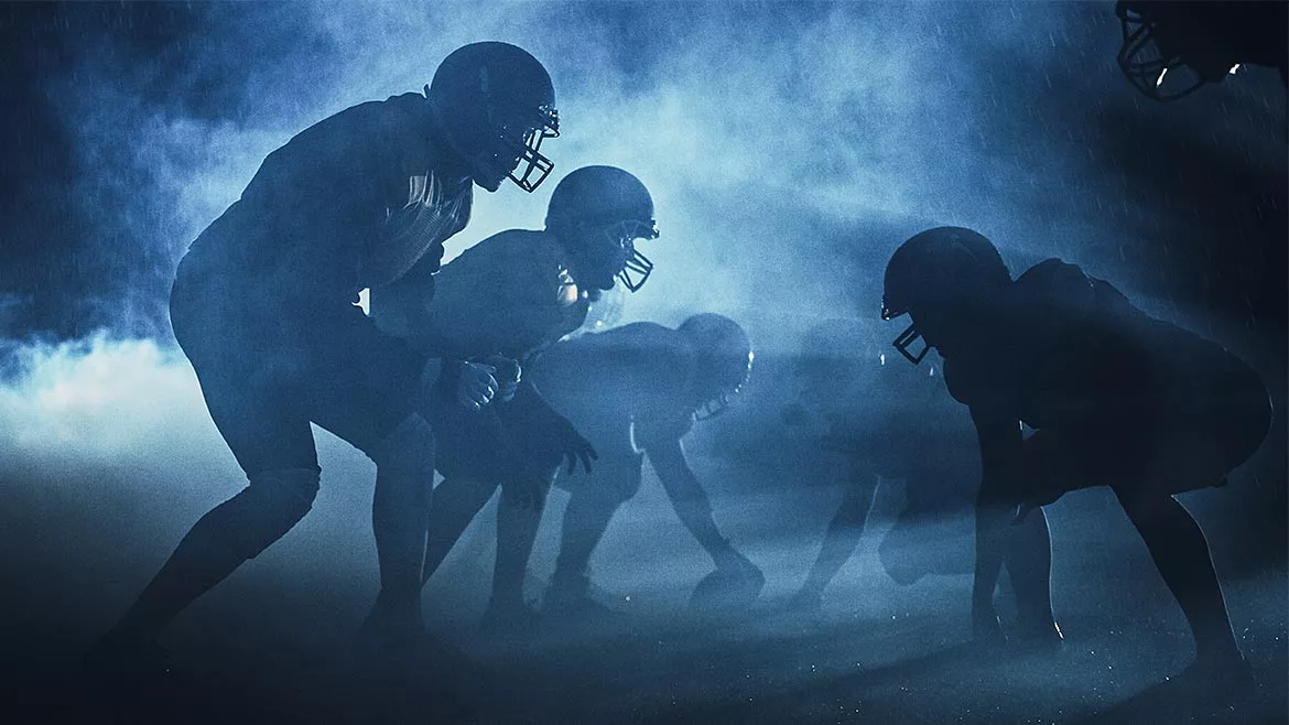 Football players on the field on a rainy, smoky night. The players are in silhouette.