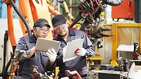 Two engineers in a factory having a discussion and looking at a tablet and clipboard.