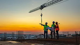 Speaking of Quality column feature image three construction workers at construction site with crane in the background.