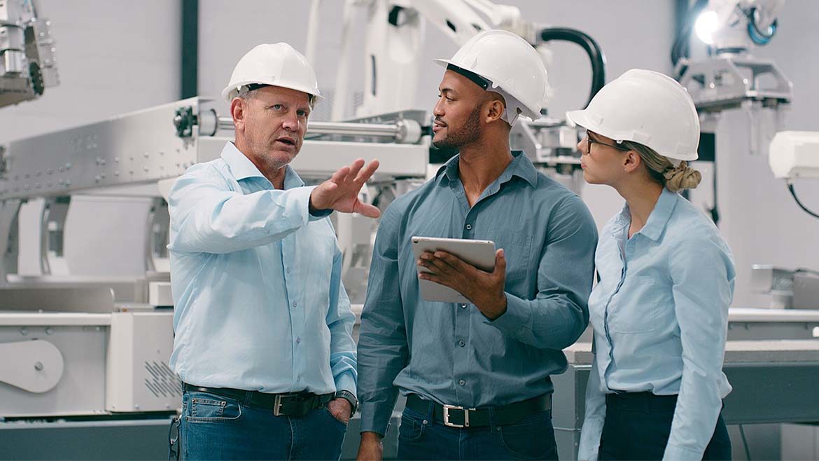 Three workers speaking, the person in the middle holding a tablet with robots in the background.