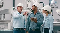 Three workers speaking, the person in the middle holding a tablet with robots in the background.