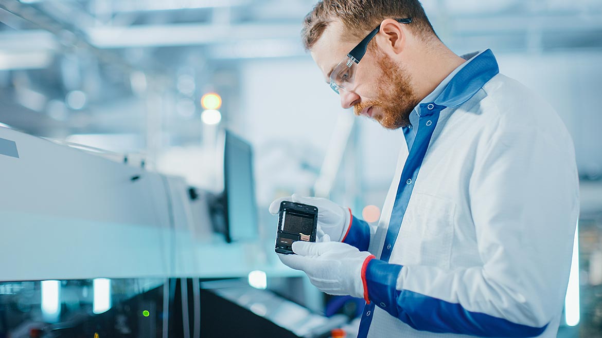 High-Tech Factory: Quality Control Engineer Picks up and Visually Checks Smart Electronic Device for Possible Damages. In the Background Assembly Line for PCB with Surface Mount Pick and Place Technology. 