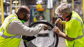 Two men working in a plastics factory. The manager, holding a digital tablet and talking to the other man, is measuring a large plastic cylinder on a conveyor belt.