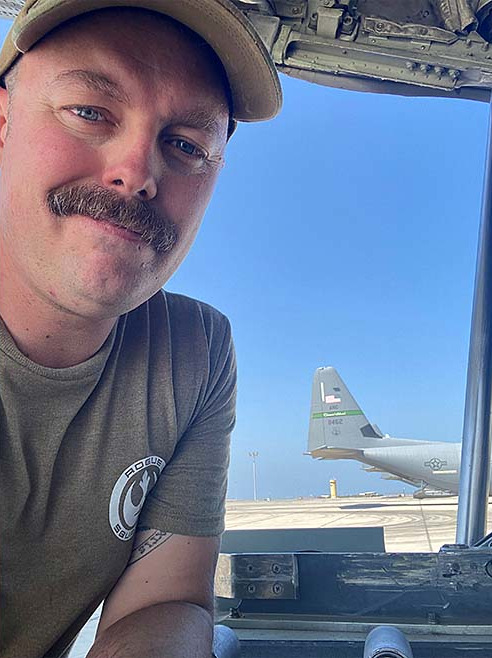 Justin Wise wearing a cap and sporting a mustache with a partial view of a military aircraft in the background.