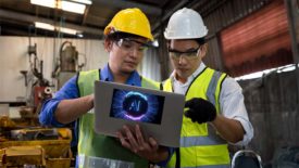 Two workers in a factory on a laptop that has "AI" image overlay.