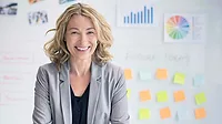 Smiling businesswoman standing in front of a whiteboard with charts and sticky notes.