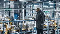 Car Factory Engineer in Work Uniform Using Tablet Computer in an Automotive Industrial Manufacturing Facility Working on Vehicle Production with Robotic Arms Technology.