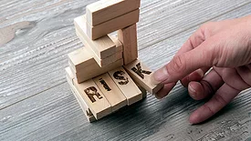 Hand taking the "K" block from wooden blocks tower. The "K" is part of four long blocks that say "RISK".