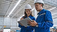 A young Asian man and woman engineer work together in a factory warehouse, using a digital tablet.