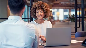 A woman using a laptop is talking with a man, his back to us, in a business setting, with a blurred background.