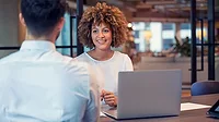 A woman using a laptop is talking with a man, his back to us, in a business setting, with a blurred background.