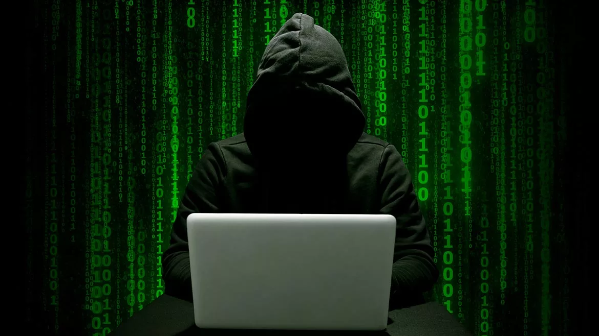 Hacker in a dark hoodie committing cyber crime with a laptop, green binary numbers on a black background.