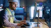 Male engineer wearing a safety vest and hard hat working on a desktop with a blurred background of the workplace.