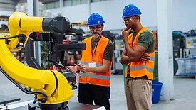 Two engineers wearing orange safety vests, safety glasses and blue hard hats, analyzing robotic arm performance in a factory setting.