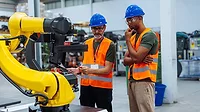 Two engineers in safety gear analyzing robotic arm performance in factory setting Two engineers wearing orange safety vests, safety glasses and blue hard hats, analyzing robotic arm performance in a factory setting.