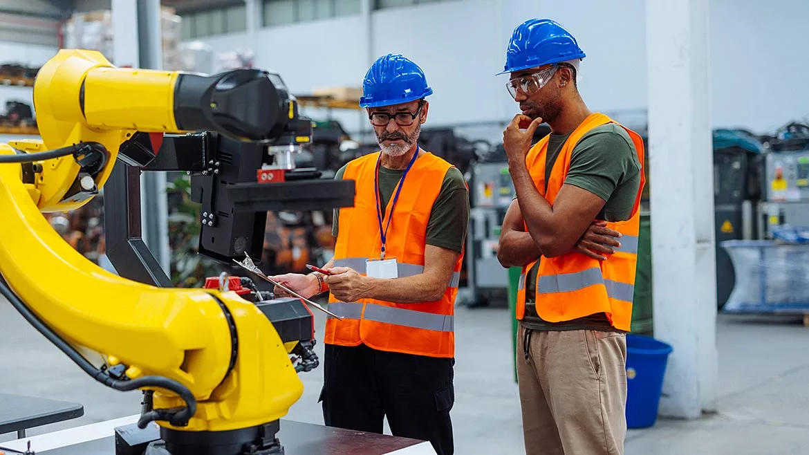 Two engineers wearing orange safety vests, safety glasses and blue hard hats, analyzing robotic arm performance in a factory setting.