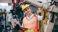 Female industrial worker in hard helmet and uniform checking on robotic arm Female industrial engineer or technician worker in hard helmet and uniform using laptop checking on robotic arm machine.