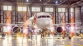 Large-scale inspection of all aircraft systems in the aircraft hangar by worker mechanics and other specialists. Bright light streaming outside from the garage door.