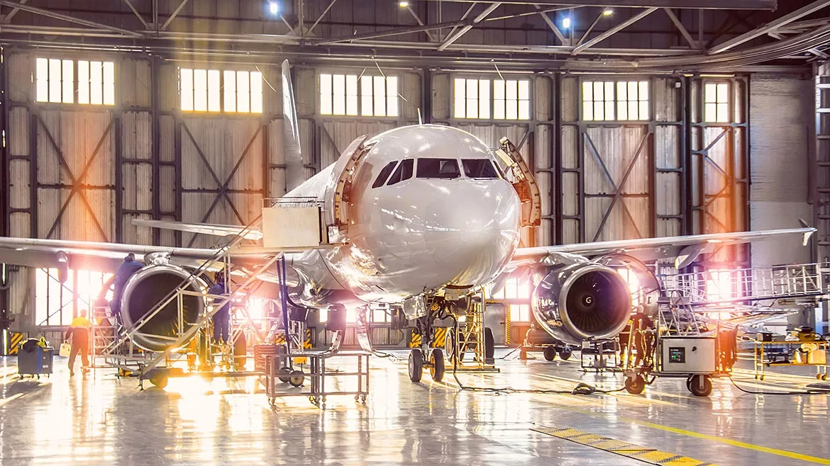 Large-scale inspection of all aircraft systems in the aircraft hangar by worker mechanics and other specialists. Bright light streaming outside from the garage door.