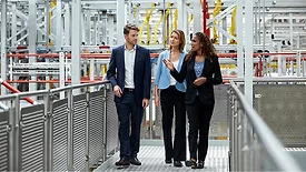 One male and two female coworkers are walking on the walkway in the factory. They are wearing business wear.