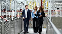 One male and two female coworkers are walking on the walkway in the factory. They are wearing business wear.