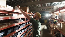 Worker picking up a small box from the top shelf at a manufacturing warehouse.