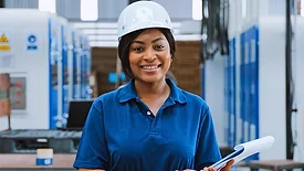 Portrait of a black female engineer technician smiling, wearing a hard hat and holding a clipboard in a warehouse packaging factory.