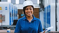 Portrait of a black female engineer technician smiling, wearing a hard hat and holding a clipboard in a warehouse packaging factory.